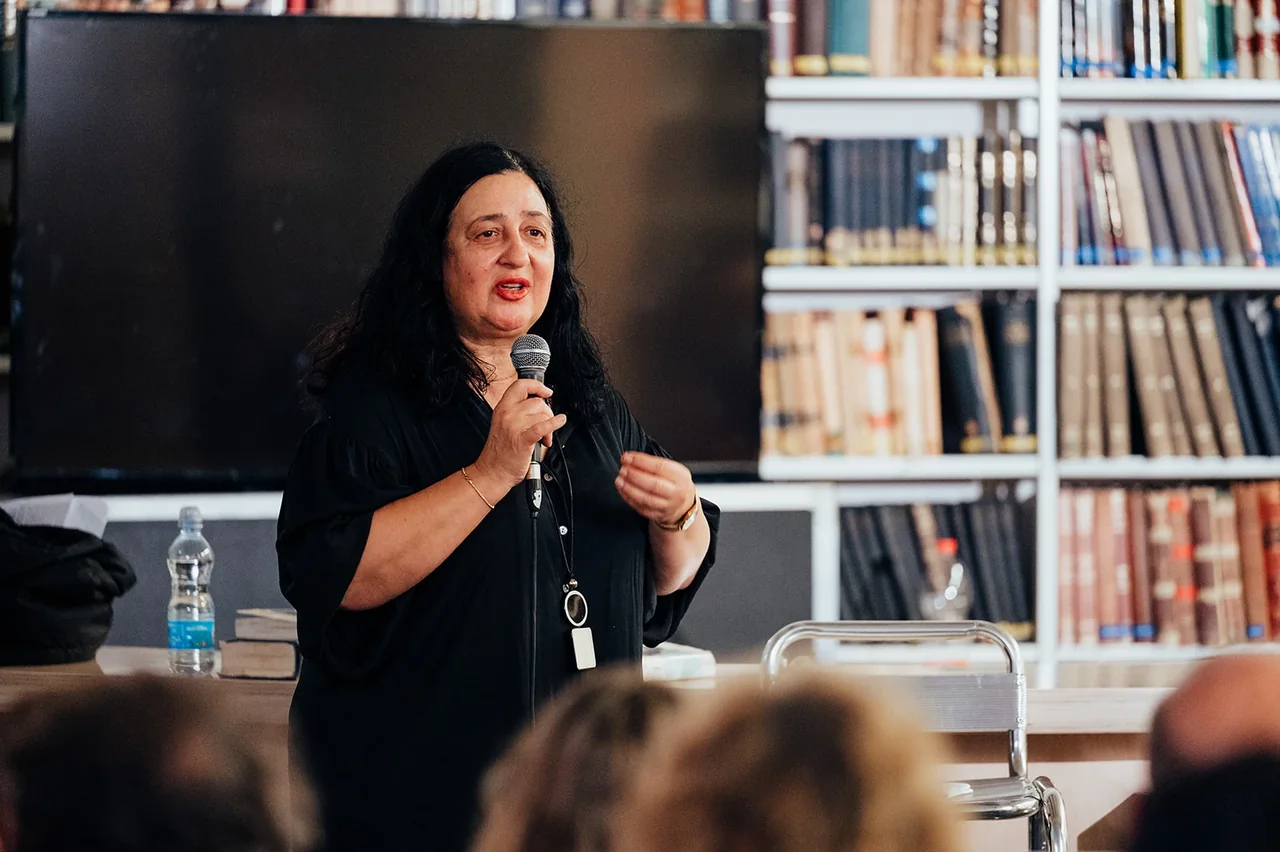 דוברת מול הקהל לצד מדפי הספרייה — צילום אמנון חורש